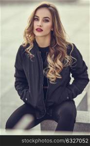 Young blonde girl with beautiful blue eyes wearing black jacket and trousers outdoors. Pretty russian female with long wavy hair hairstyle. Woman in urban background.