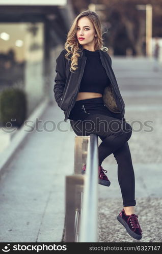 Young blonde girl with beautiful blue eyes wearing black jacket and trousers outdoors. Pretty russian female with long wavy hair hairstyle. Woman in urban background.