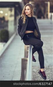 Young blonde girl with beautiful blue eyes wearing black jacket and trousers outdoors. Pretty russian female with long wavy hair hairstyle. Woman in urban background.