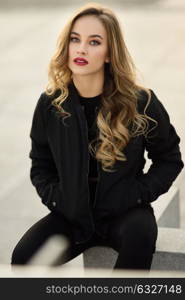 Young blonde girl with beautiful blue eyes wearing black jacket and trousers outdoors. Pretty russian female with long wavy hair hairstyle. Woman in urban background.