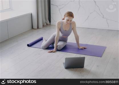 Young blonde happy woman practicing yoga on mat. White girl is watching video on laptop or talking to coach. Concept of e-learning and home online classes using computer. Video workout.. Young happy white woman practicing yoga watching video on laptop. Concept of e-learning.