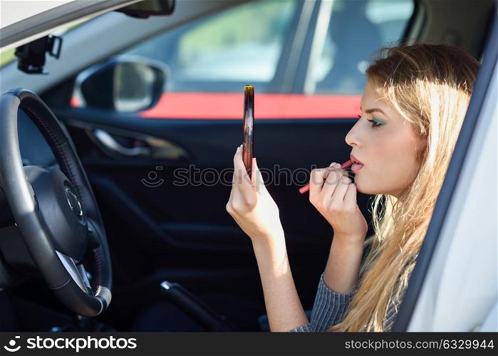 Young blonde woman applying lipstick looking at mirror in her car. Girl making-up herself in urban background.