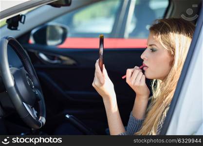 Young blonde woman applying lipstick looking at mirror in her car. Girl making-up herself in urban background.