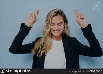 Young blonde woman in black blazer holding both hands in air with crossed fingers, crossing them with hope and wishing for good luck while posing isolated over blue studio background. Young blonde woman making wish with crossed fingers