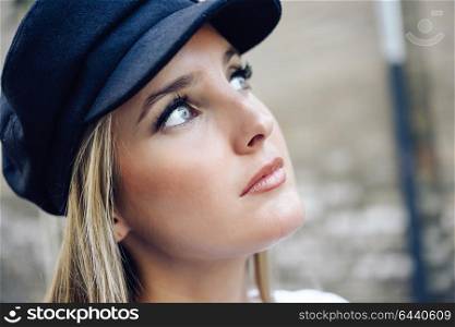 Young blonde woman in defocused urban background. Girl with blue eyes wearing cap.
