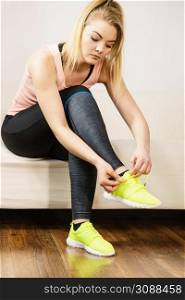 Young blonde woman in sportswear sitting on sofa indoor getting ready for exercises. Tying up shoes. Woman putting sport shoes