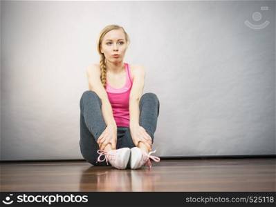 Young blonde woman in sportswear sitting on wooden floor indoor getting ready for exercises. Training at home, being fit and healthy.. Woman in sportswear sitting on ground