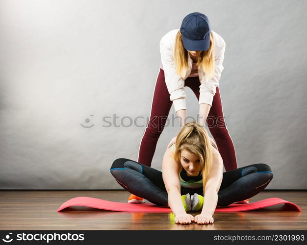 Young blonde woman in sportswear sitting on wooden floor indoor stretching legs with her female trainer. Training at home, being fit and healthy.. Woman in sportswear stretching legs with trainer