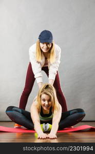 Young blonde woman in sportswear sitting on wooden floor indoor stretching legs with her female trainer. Training at home, being fit and healthy.. Woman in sportswear stretching legs with trainer