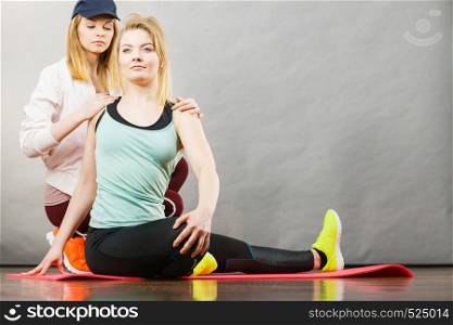 Young blonde woman in sportswear sitting on wooden floor indoor stretching legs with her female trainer. Training at home, being fit and healthy.. Woman in sportswear stretching legs with trainer