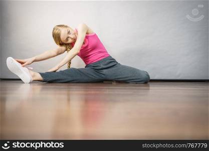 Young blonde woman in sportswear sitting on wooden floor indoor stretching legs. Training at home, being fit and healthy.. Woman in sportswear stretching legs