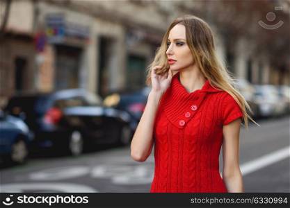 Young blonde woman looking at something in the street. Beautiful girl in urban background wearing red dress. Female with straight hair.