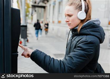 Young blonde woman paying on parking meter in the city with surprised face wearing a jacket an headphones