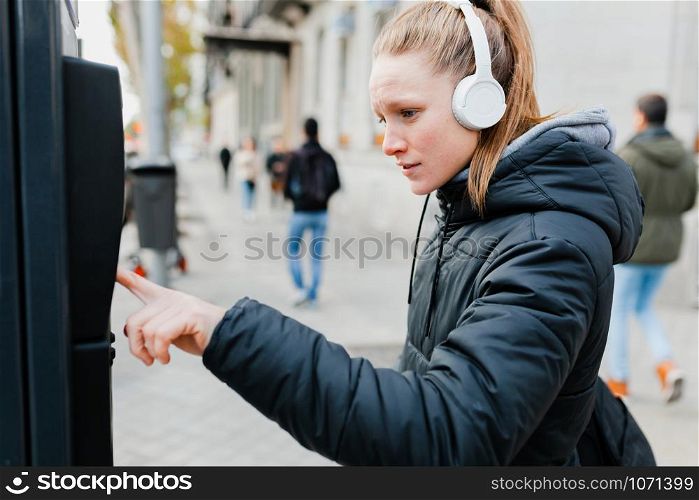 Young blonde woman paying on parking meter in the city with surprised face wearing a jacket an headphones