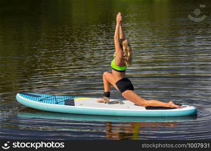 Young blonde woman practicing warrior 1 posture on SUP at water surface