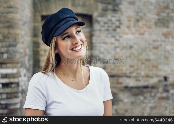 Young blonde woman smiling near a brick wall. Girl with blue eyes wearing white t-shirt and cap.