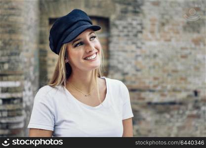 Young blonde woman smiling near a brick wall. Girl with blue eyes wearing white t-shirt and cap.