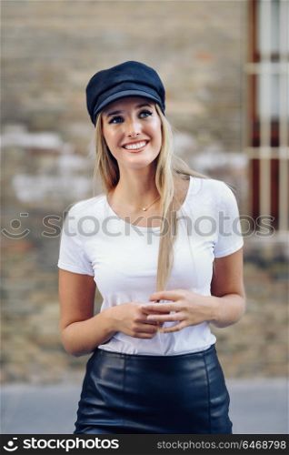 Young blonde woman smiling near a brick wall. Girl with blue eyes wearing t-shirt, skirt and cap.