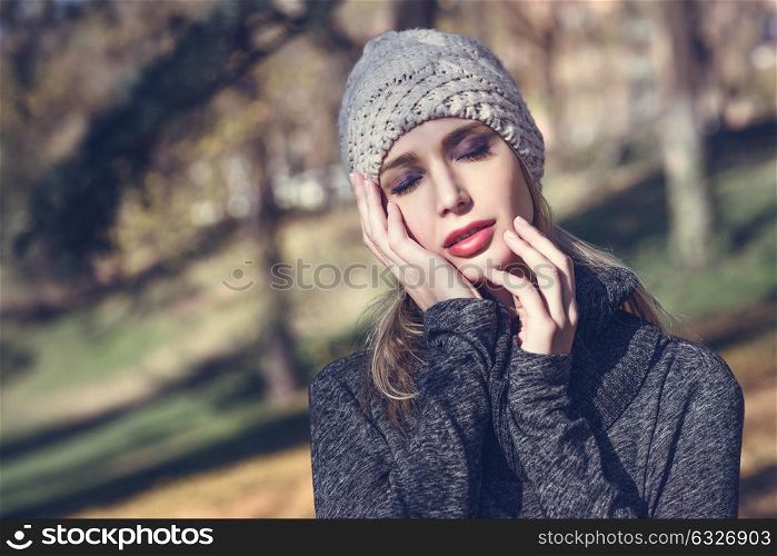 Young blonde woman standing in a park with autumn colors. Beautiful girl wearing winter gray dress and wool cap. Female with straight hair and eyes closed with dark eyeshadow.