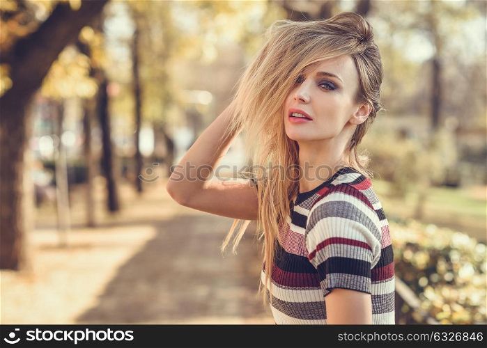 Young blonde woman standing in the street moving her hair. Beautiful girl in urban background wearing striped dress. Female with straight hair and blue eyes.