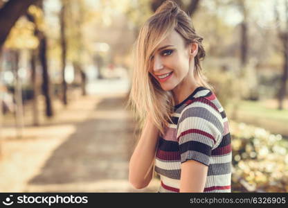 Young blonde woman standing in the street moving her hair. Beautiful girl in urban background wearing striped dress. Female with straight hair and blue eyes.