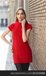 Young blonde woman standing in the street near a brick wall. Beautiful girl in urban background wearing red dress and black tights. Female with straight hair.