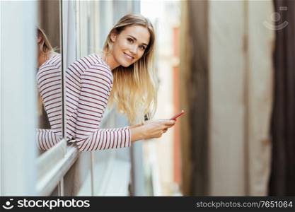 Young blonde woman using a smartphone looking at camera. Girl leaning out of her house window.. Young woman leaning out of her house window using a smartphone.