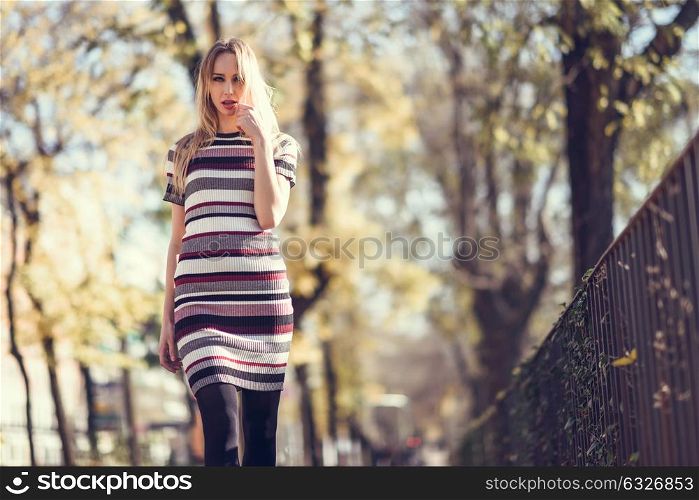Young blonde woman walking in the street. Beautiful girl in urban background wearing striped dress and black tights. Female with straight hair.