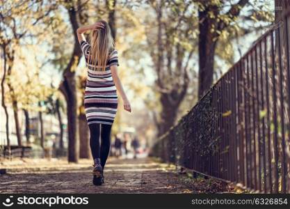 Young blonde woman walking in the street. Beautiful girl in urban background wearing striped dress and black tights. Female with straight hair. Back view