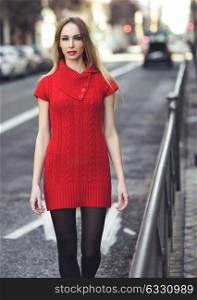 Young blonde woman walking in the street. Beautiful girl in urban background wearing red dress and black tights. Female with straight hair.