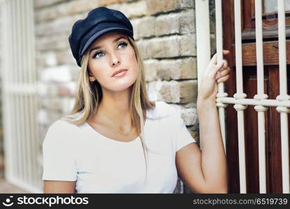Young blonde woman wearing cap standing near a brick wall.. Young blonde woman standing near a brick wall. Girl with blue eyes wearing white t-shirt and cap.