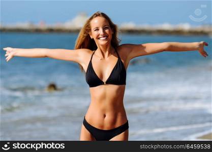 Young blonde woman with beautiful body in swimwear on a tropical beach with open arms. Caucasian female with straight long hairstyle wearing black bikini.