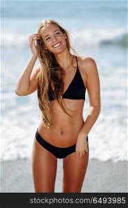 Young blonde woman with beautiful body in swimwear smiling on a . Young blonde woman with beautiful body in swimwear smiling on a tropical beach. Caucasian female with straight long hairstyle wearing black bikini.