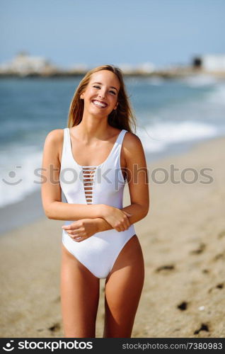 Young blonde woman with beautiful body in white swimsuit on a tropical beach. Caucasian female with straight long hairstyle smiling.. Blond woman with beautiful body in swimswit on a tropical beach