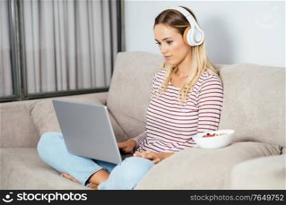 Young blonde woman with headphones and laptop on the sofa at home.. Young blonde woman with headphones and laptop on the sofa