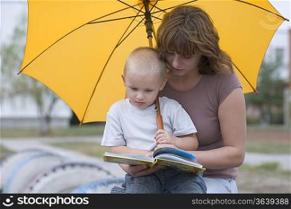 Young boy and woman sit under yellow umbrella reading a book