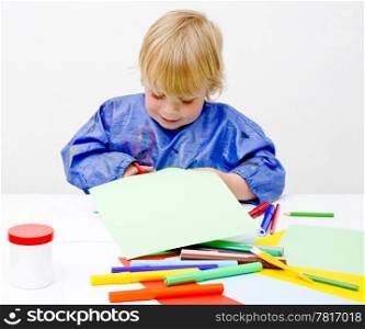 Young boy happily cutting his way into a sheet of paper on a table filled with colorful pencils, glue en felt pens