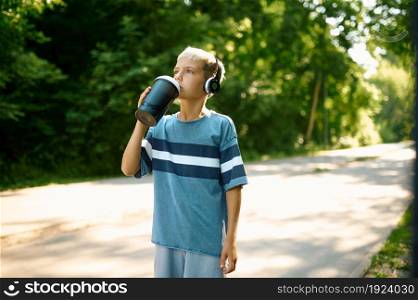 Young boy in headphones drinks water on walking path outdoors. Child in earphones wakling in summer park, kid is thirsty. Young boy in headphones drinks water outdoors