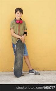 Young boy leaning on yellow wall with headphones on neck, holding a skateboard while looking away in a bright day