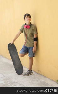 Young boy leaning on yellow wall with headphones on neck, holding a skateboard while looking away in a bright day