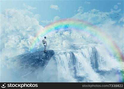 Young boy on a cliff looking at the horizon with a waterfall above clouds. Life journey below a rainbow in paradise. Inspirational imaginary view.