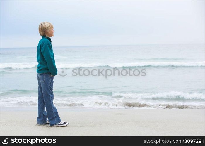 Young Boy On Holiday Standing On Winter Beach Looking Out To Sea