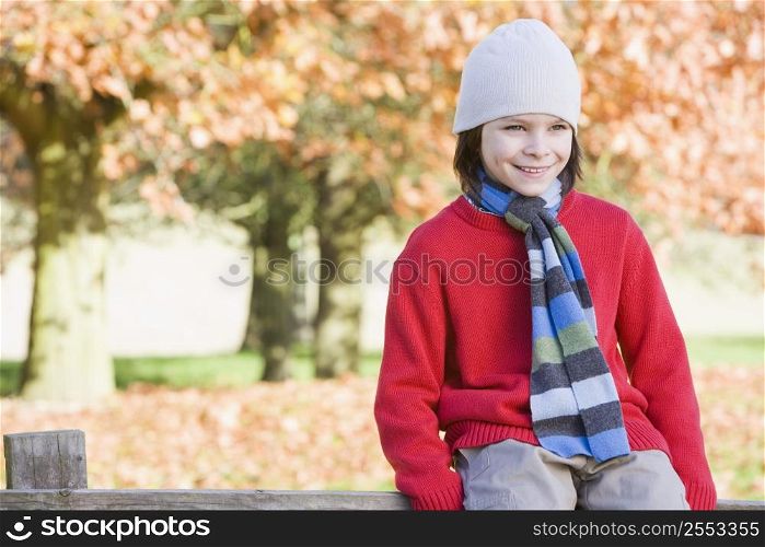 Young boy outdoors at park sitting on fence smiling (selective focus)