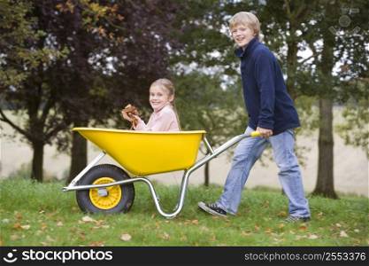Young boy outdoors pushing young girl in wheelbarrow and smiling (selective focus)