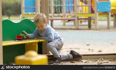 Young boy playing outdoors in a childrens playground crouching down at a colourful little table playing with toy tractor