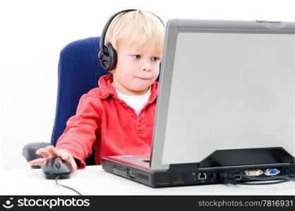 Young boy sitting behind a laptop, clicking on a computermouse, whilst looking at the screen and wearing headphones