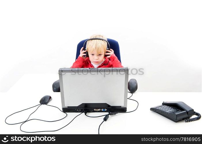 Young boy, sitting behind a laptop, wearing a head set, looking at the screen with a phone and a computer mouse next to him