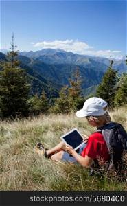Young boy sitting in a mountain meadow using a tablet computer. Summer season, clear blue sky. Boy is wearing a red shirt, white cap and a grey backpack. Piemonte, West Italian Alps.