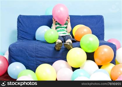 Young boy sitting on a couch, hiding behind a baloon he&rsquo;s holding
