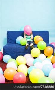 Young boy sitting on a couch, hiding behind a pink baloon he&rsquo;s holding